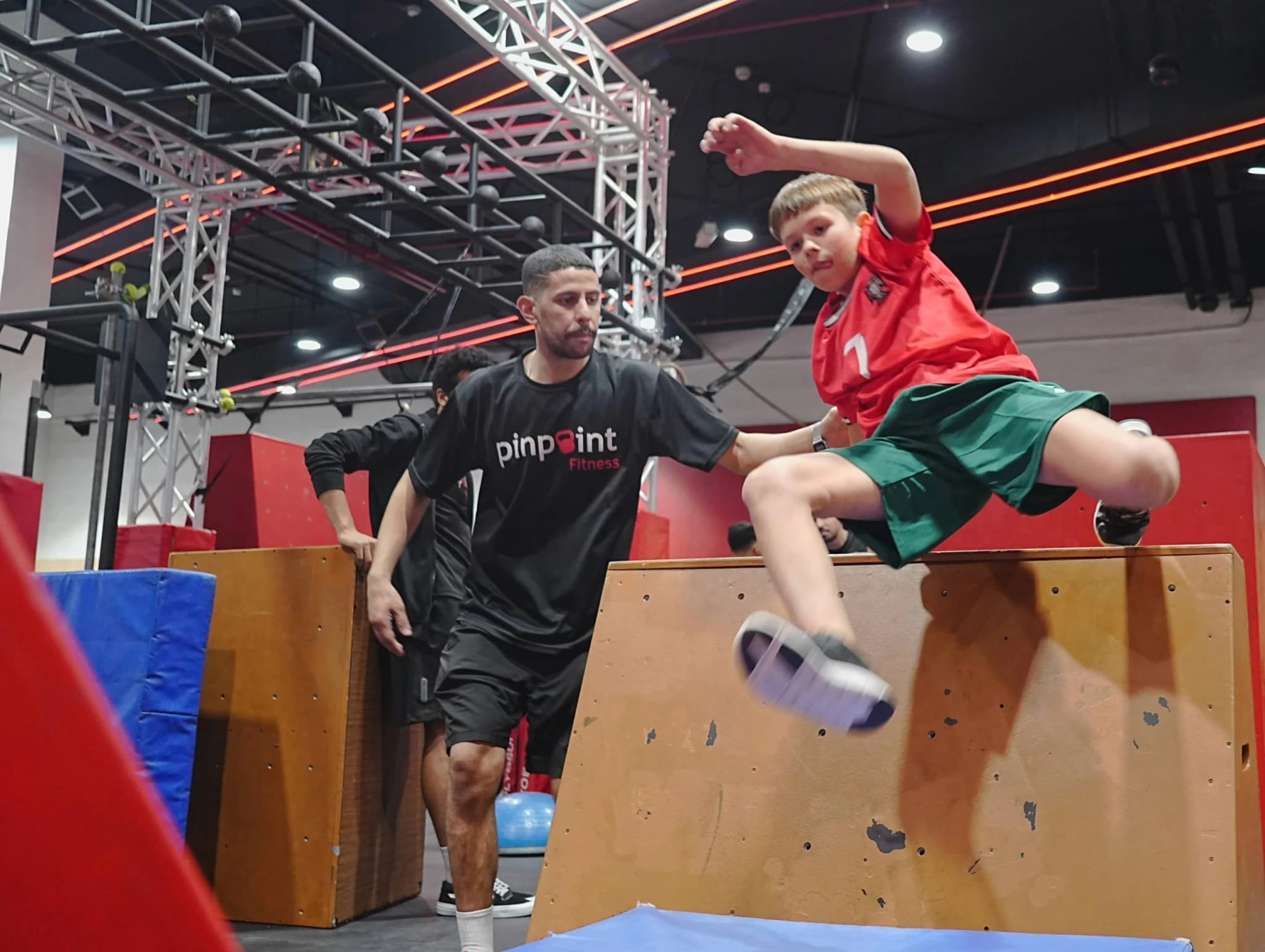 A boy leaping over an obstacle box as his Pinpoint Fitness coach spots him